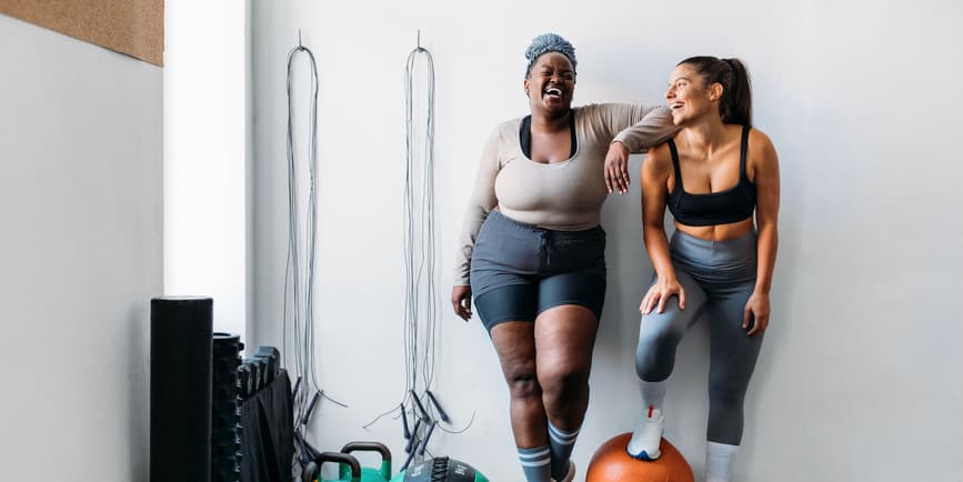 Una mujer negra con curvas vestida con ropa deportiva y un pañuelo para el cabello y una mujer blanca musculosa vestida con ropa deportiva están juntas, riendo, descansando junto al pozo de un gimnasio.