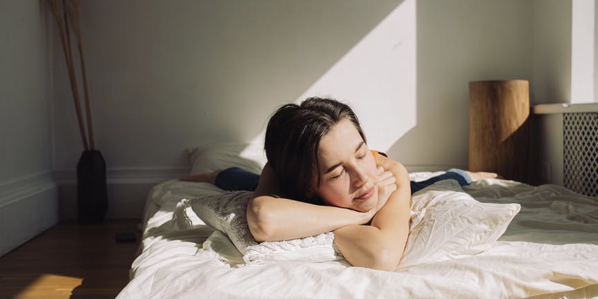 Mujer joven acostada en un colchón en el suelo a la luz del sol con los ojos cerrados