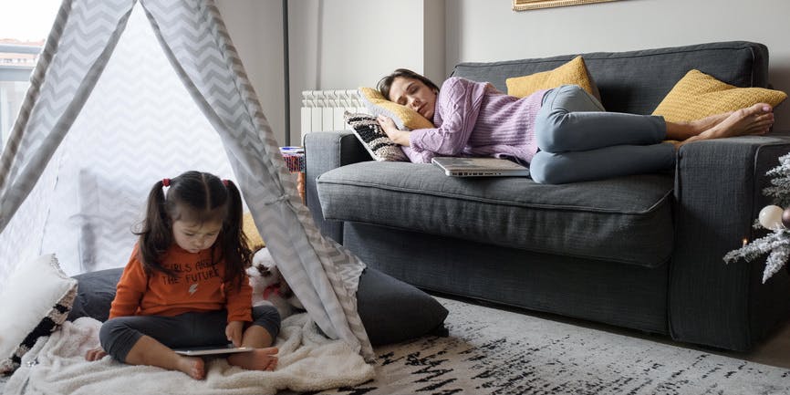 Fotografía en color de una madre joven echándose una siesta en el sofá de la sala de estar mientras su hija pequeña lee un libro dentro de una carpa de gasa.