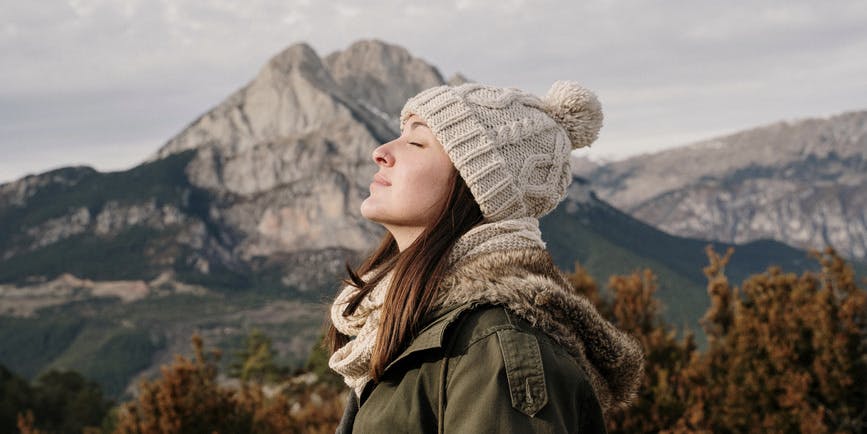 Una joven blanca se relaja con los ojos cerrados frente a un paisaje pintoresco, vestida con ropa de invierno y un sombrero.