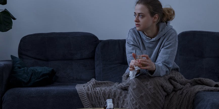 Mujer enferma tomando frío sentada con un vaso de té caliente en el sofá debajo de una manta y mirando hacia otro lado. Se está recuperando con la ayuda de un medicamento.