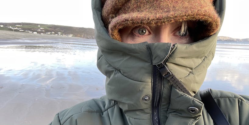 Una selfie de una mujer en una playa en invierno. Está envuelta contra el frío, con un abrigo aislante con la capucha levantada y un gorro de lana. Lo único que se ve de su rostro son sus ojos.