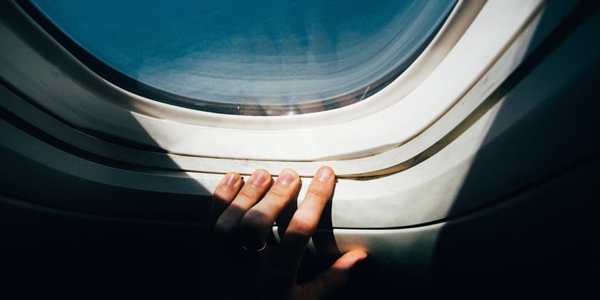 A color photograph of a hand in shadow pressing fingers against an airplane window with a blue sky and sunlight streaming in.