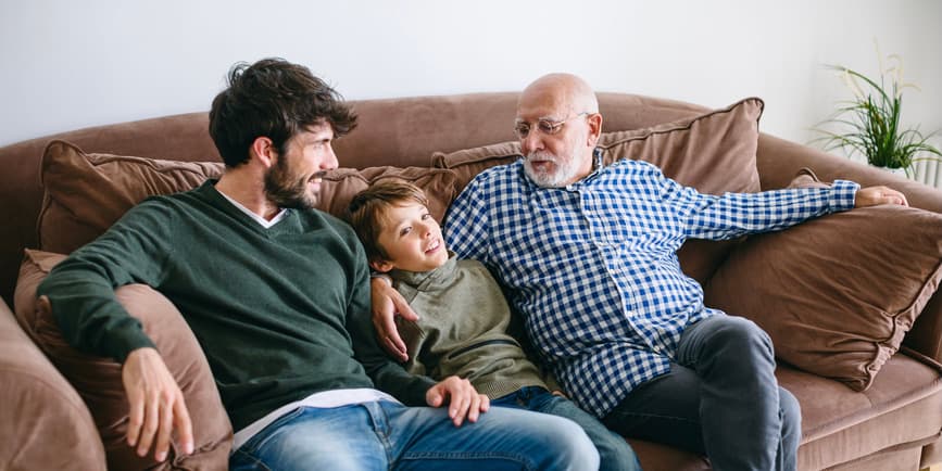 Un abuelo, un padre y un hijo en casa en el sofá, hablando y sonriendo.