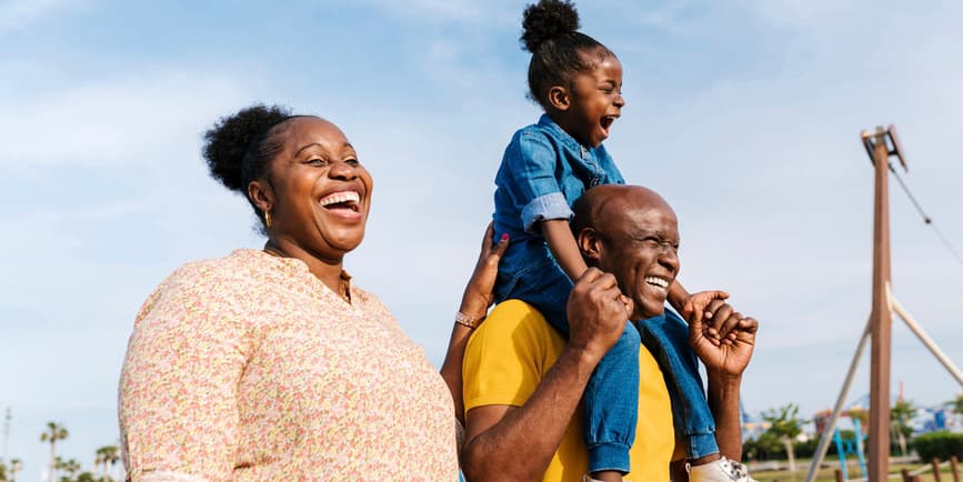 Excited black father carrying happy little daughter on shoulders while mother laughing and strolling together on playground on tropical resort