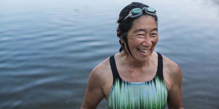 Retrato de una mujer bronceada de unos cincuenta o sesenta años que lleva un traje de baño y unas gafas, sonriendo al salir de un lago nadando.
