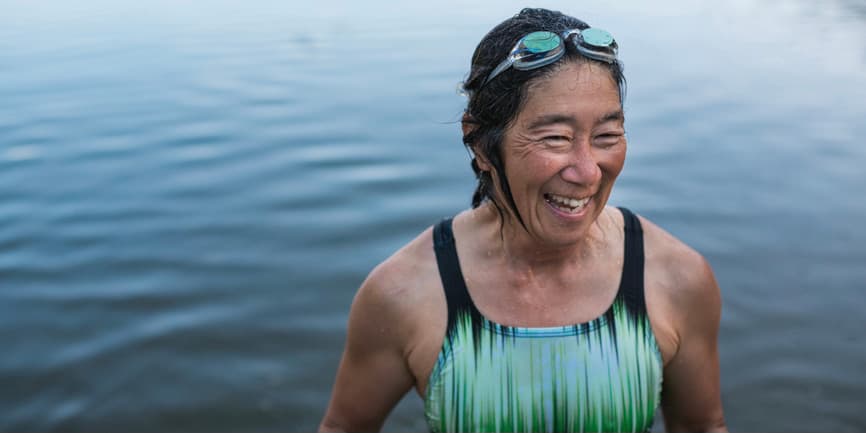 Portrait of a tanned woman in her fifties or sixties wearing a swimsuit and goggles, smiling as she comes out of swimming in a lake.