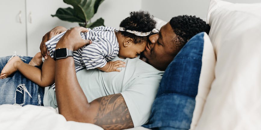 A black father wearing jeans and t-shirt lays on his bed smiling down with his infant daughter who is cuddling on top of him.