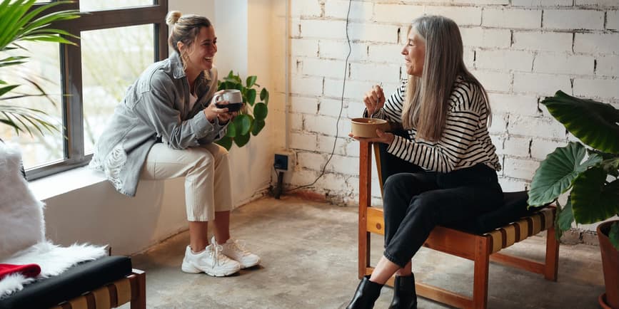 Two stylish white female colleagues, one in her twenties and one in her fifties, take a break in their modern office to have lunch and drink coffee.