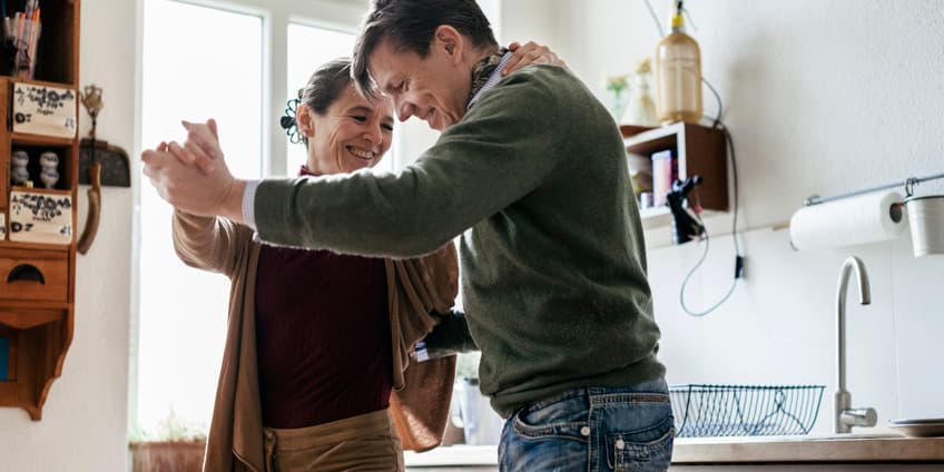 A white couple in their late forties or early fifties wearing pants and sweaters dance around in their home kitchen, smiling.