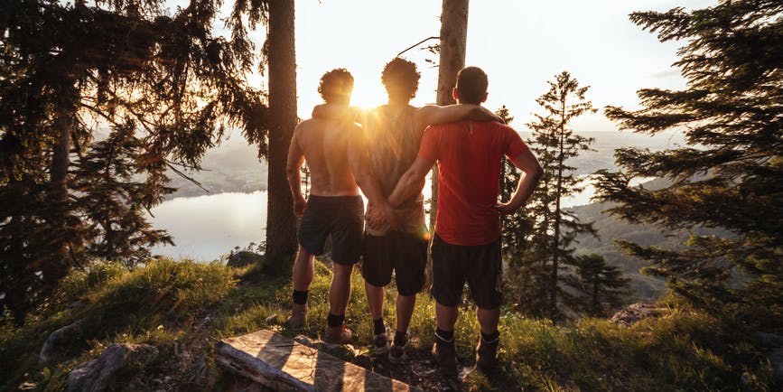 A color photo at sunset, where three buff men in hiking exercise clothes stand looking looking at the sunset over the water, their arms around and on each other.