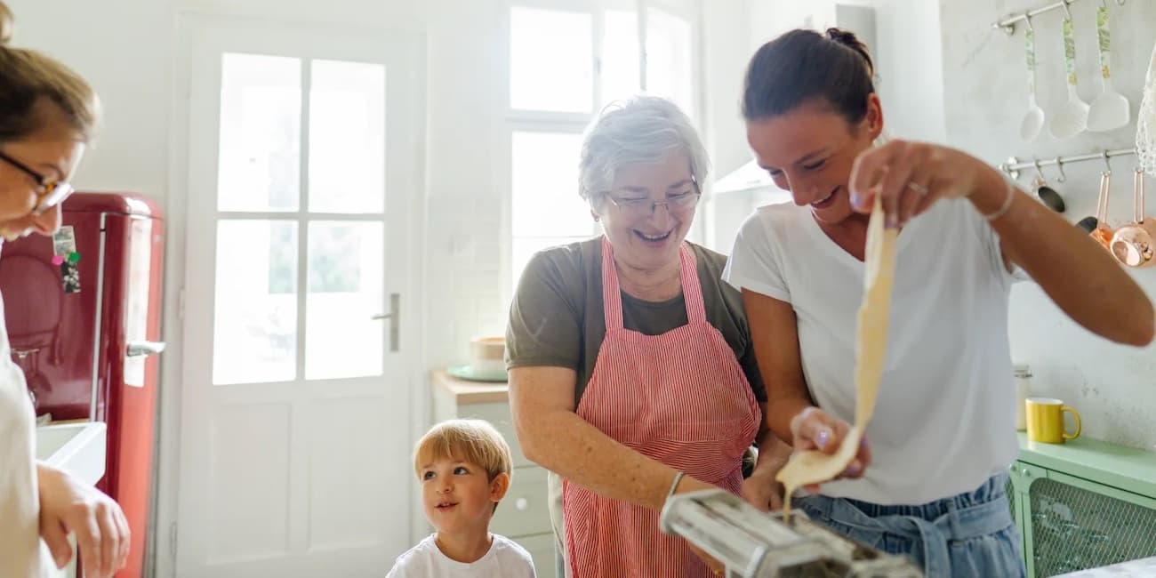 Color photograph of three generations of a white Italian family making pasta together in a sunlit kitchen.