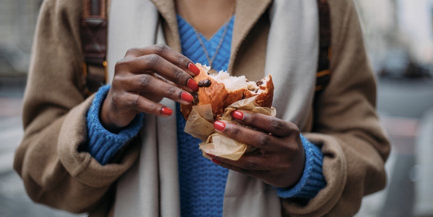 Outside closeup photograph of the hands of a young Black woman with red fingernails wearing a blue sweater and tan coat eating a bready pastry.