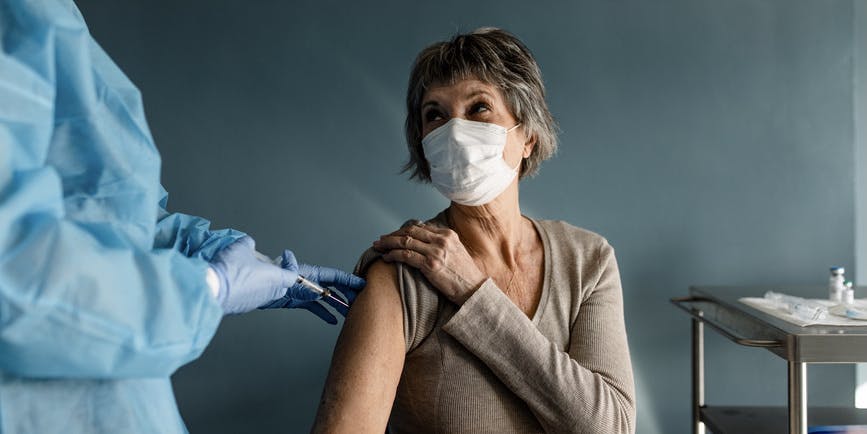 An attractive senior woman with short silver hair wearing a longsleeved tan shirt and white medical mask gets a Covid-19 injection in a blue-walled office by a practitioner wearing a medical gown, gloves and mask.