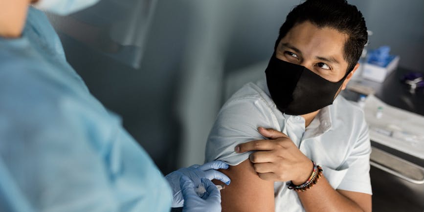A young Latino man wearing a shortsleeved button down shirt and black 95 mask gets a Covid-19 vaccine in a medical office by a gowned practitioner wearing gloves and a medical mask.