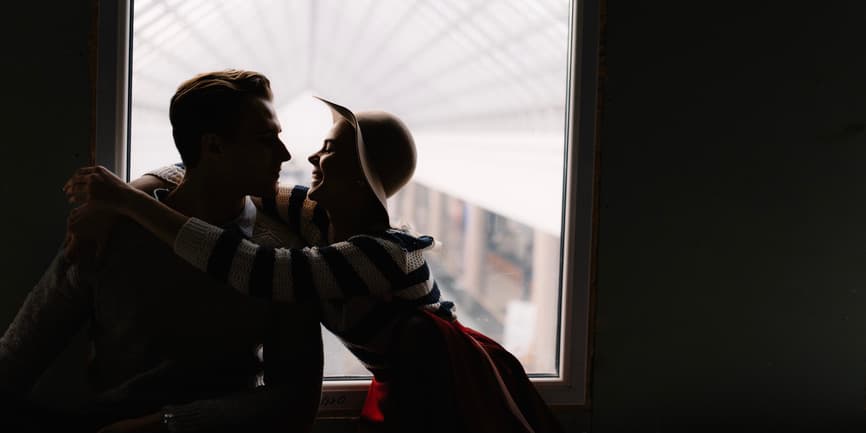 Side view couple in love enjoy to be together, woman hugging man while saying romantic words in dark room in front of window