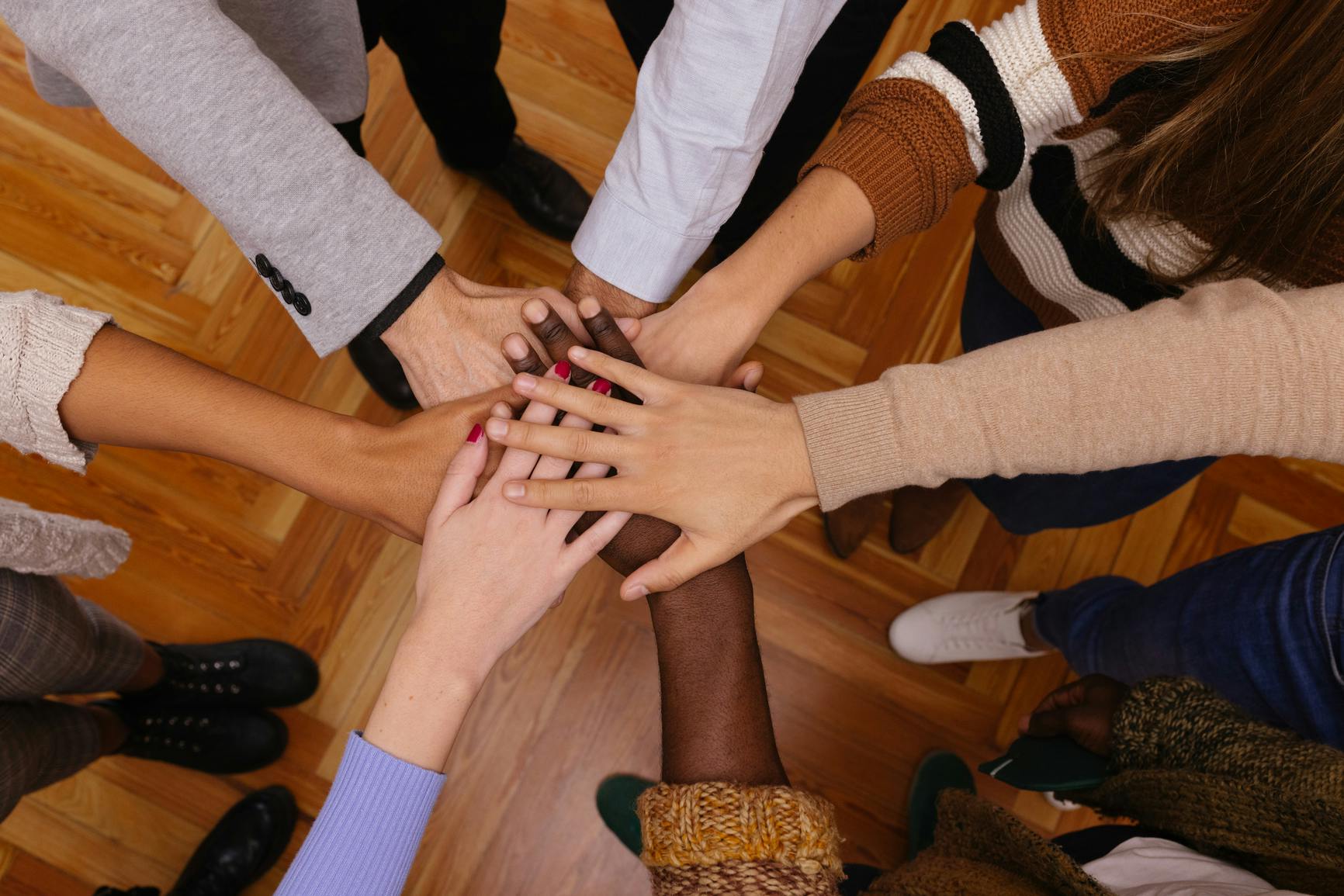 Fotografía en primer plano de un grupo de personas de pie con los brazos apuntando hacia adentro, formando un círculo con las manos superpuestas.