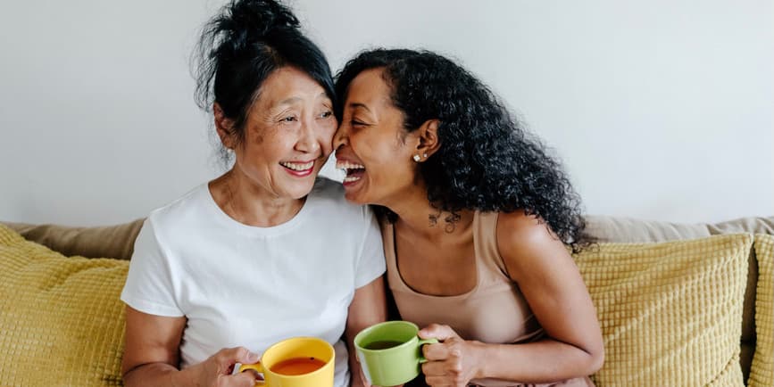 Senior asian mother with laughing adult daughter talking while drinking tea sitting on couch at home