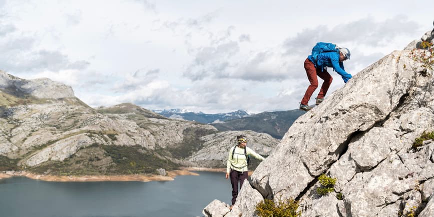 A color photo of a mountainous landscape where a man and woman in their fifties wearing hiking clothes scale a large rock on a cloudy spring day. 