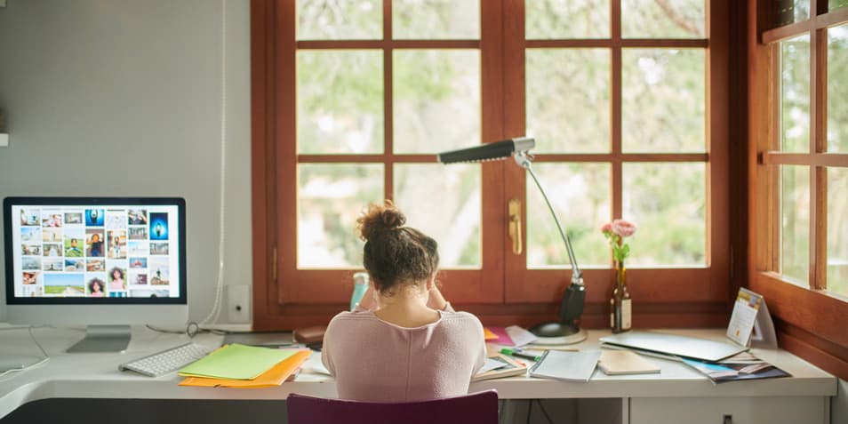 Back view of unrecognizable sad white with head down sitting at colorful workplace with notebooks scattered around, frustrated.