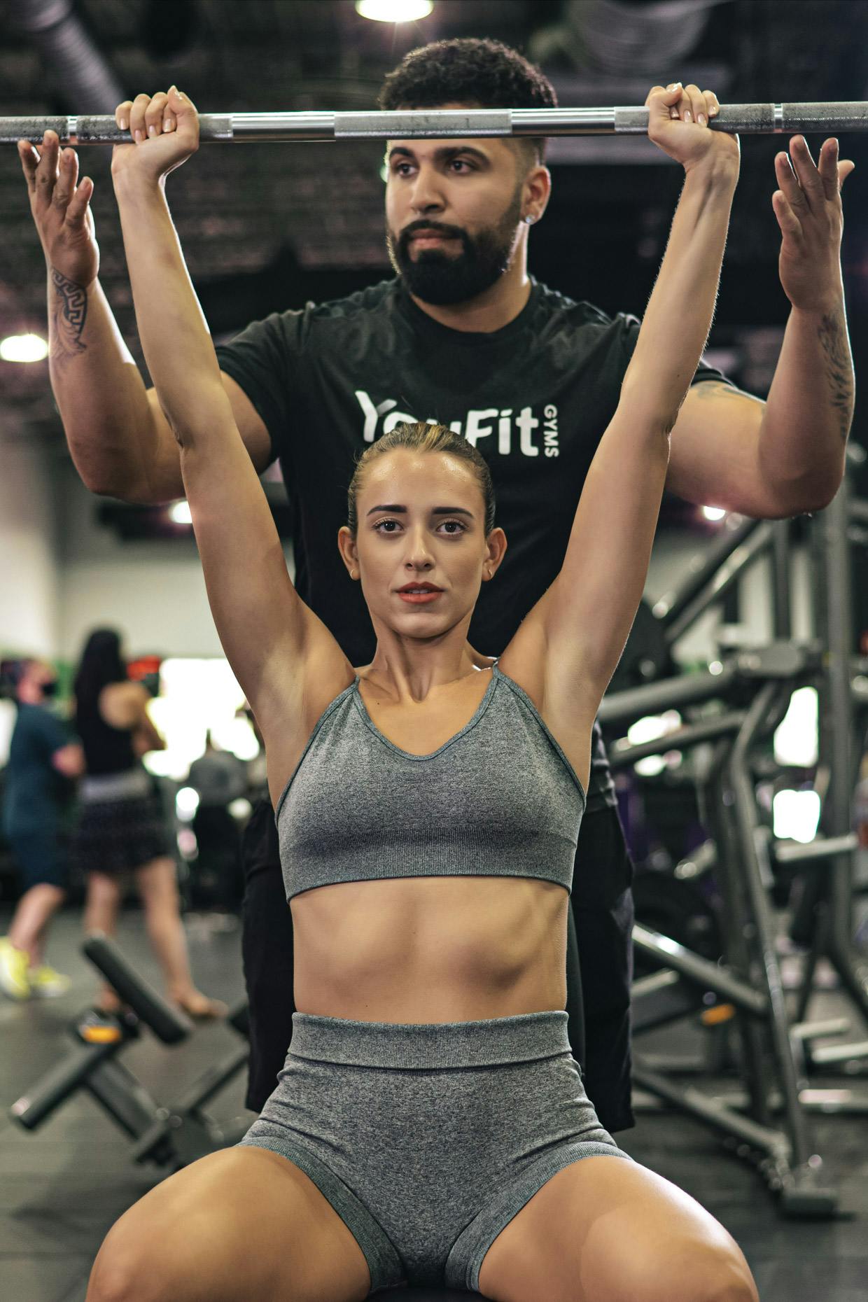A woman sitting down lifting weights in a gym being helped by a trainer