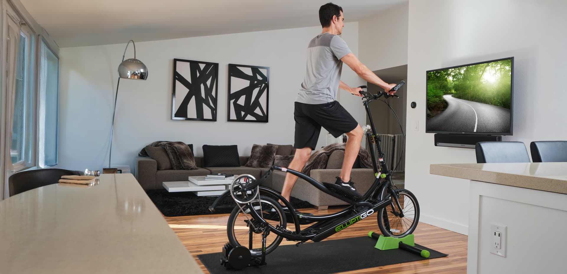 A man using an elliptical machine indoors while watching a TV screen in their livingroom