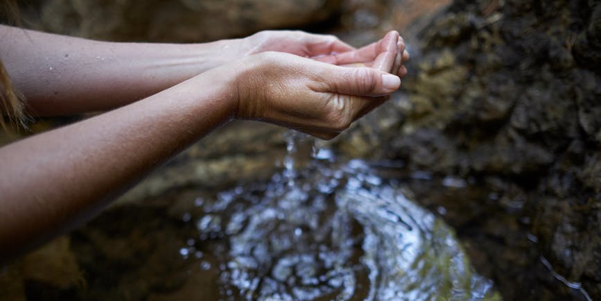 Closeup photo of a white women's arm as she holds water in her hands from a rocky stream.