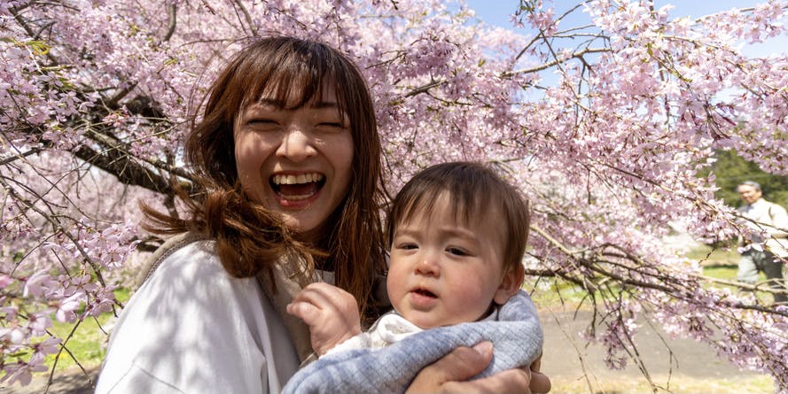 Una mujer joven de cabello castaño que lleva un jersey sonríe mientras sostiene a su hijo pequeño frente a un cerezo japonés florecido.