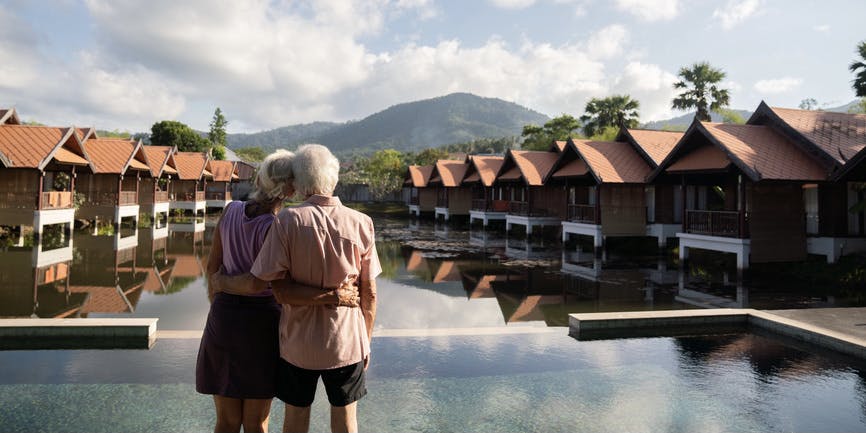 View from the back of a senior couple holding onto each other and enjoying a scenic mountain nature view overlooking a hotel pool area.