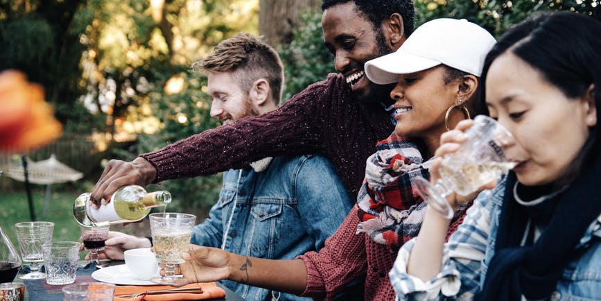An outdoor photo of a diverse, attractive group of friends dining al fresco, with a young Black male host smiling as he pours wine into a friend's glass.