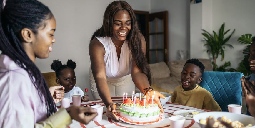 A Black woman in their living room presents a birthday cake with lit candles for her daughter. They are celebrating her birthday at home as a family with her brothers and little cousins. Everyone is happy enjoying the moment together