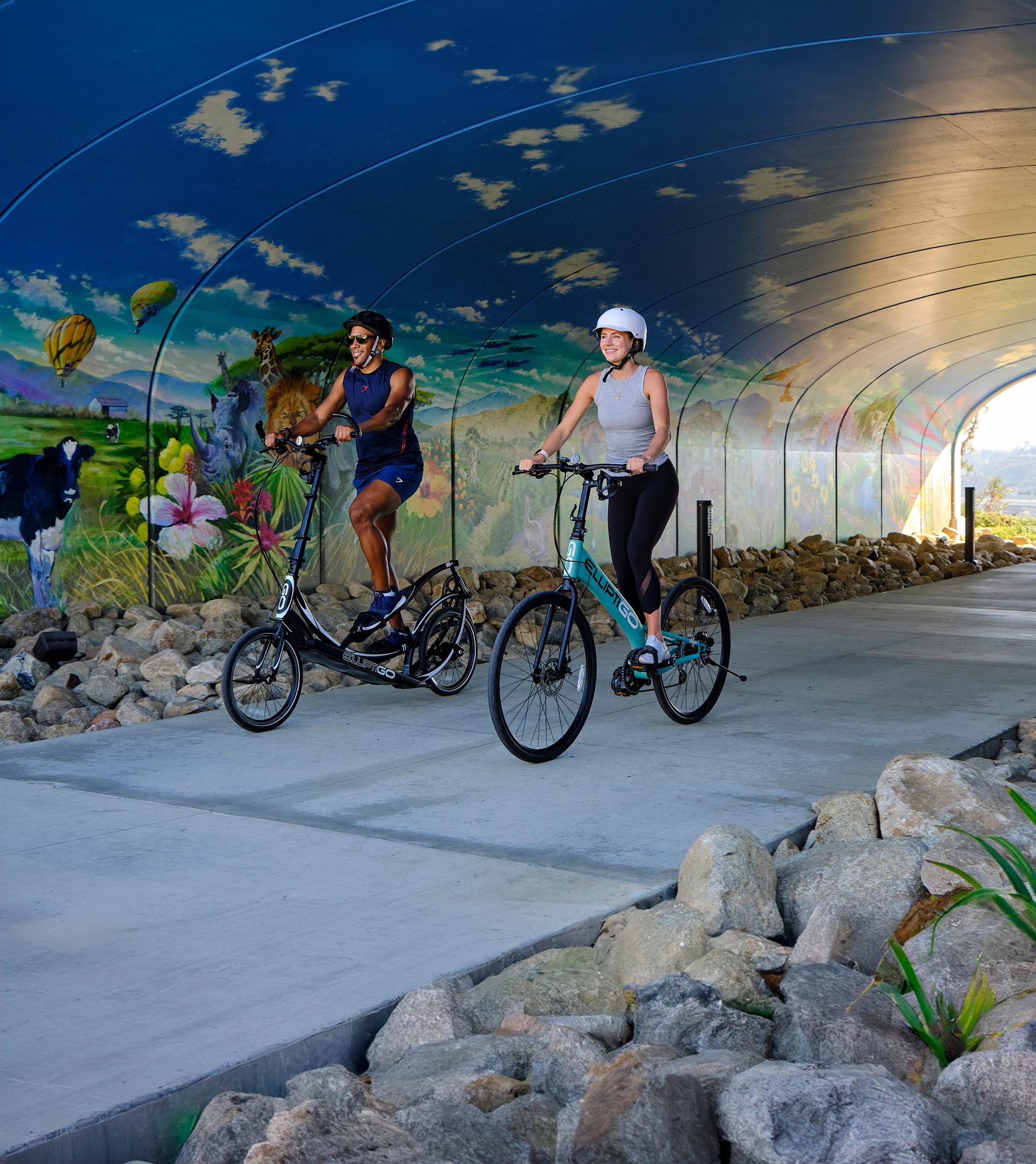 A couple biking in an elliptical bike and a standup bike going under a tunnel