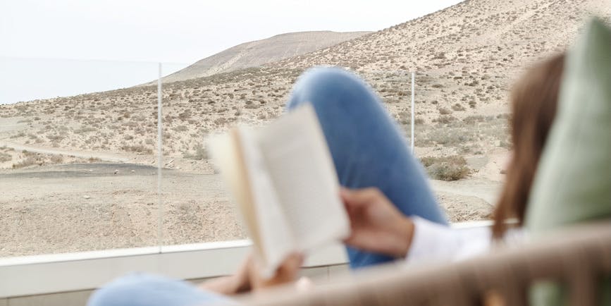 Young woman reading a book while relaxing outside on a chair on her patio with a view inside Jandía National Park overlooking the Atlantic Ocean in Sotavento Beach, Costa Calma, Fuerteventura, Canary Islands, Spain.