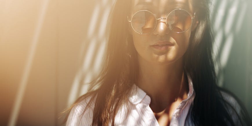 Portrait of a young woman with tanned skin and long dark hair wearing round sunglasses and a collared shirt standing in natural sunlight with shadows on her face.