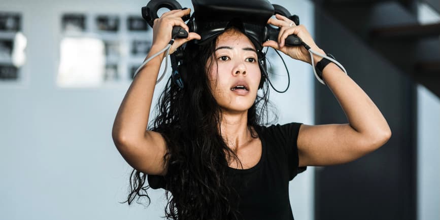 A sweaty young Asian woman with long wavy black hair wearing a black t-shirt stares at something away from the camera as she lifts a VR device from her head.