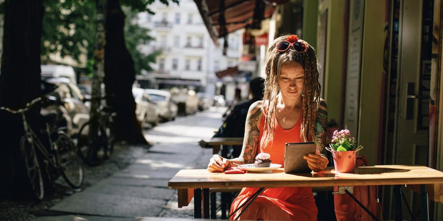 A white woman with long dreadlocks and tattoos wearing a summer dress sits at an outdoor cafe, reading an ebook.
