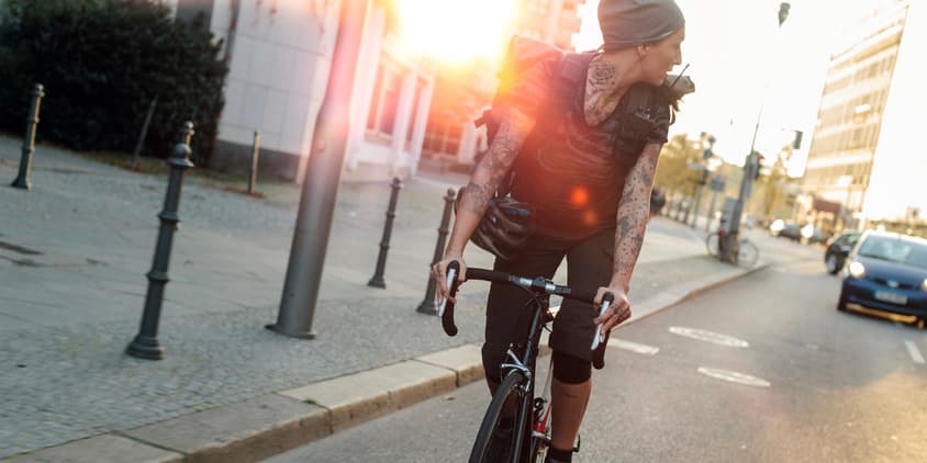 A fit white bike messenger with tattoos wearing all black clothing rides down a city street, looking over their shoulder into traffic, with the setting sun casting shadows around them.