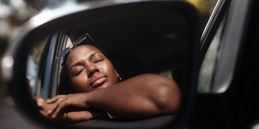 A young Black woman inside a car rests with her eyes closed against the sun and her arms on the open door window frame, seen in the side mirror. 