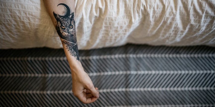 The arm of a white man with an owl tattoo leaning out from bed.