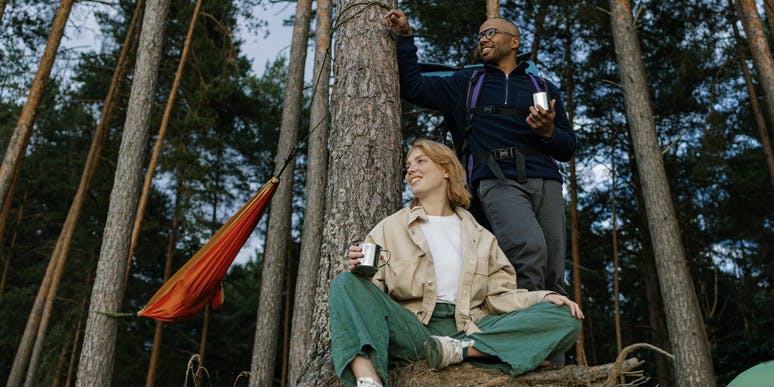 A young Black man with a bald head wearing  and a young white woman with red hair wear camping clothes and drink coffee from metal mugs at their woodland campsite, smiling as they look at something off camera.