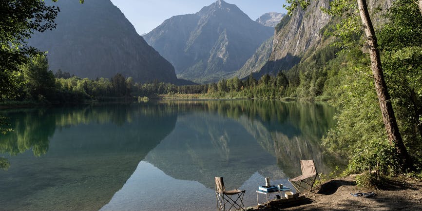 A broad view of an alpine lake in the mountains, with camping chairs, table, gas stove and coffee maker sitting at the edge and no people in sight.