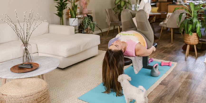 Curvy woman lying on an adjustable weight bench training from home accompanied by her cute puppy in living room