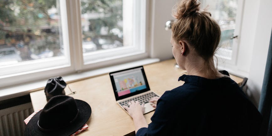 A young woman with her hair tied up, wearing a long sleeved shirt, is seen from behind, working on a laptop in front of a sunlit window.