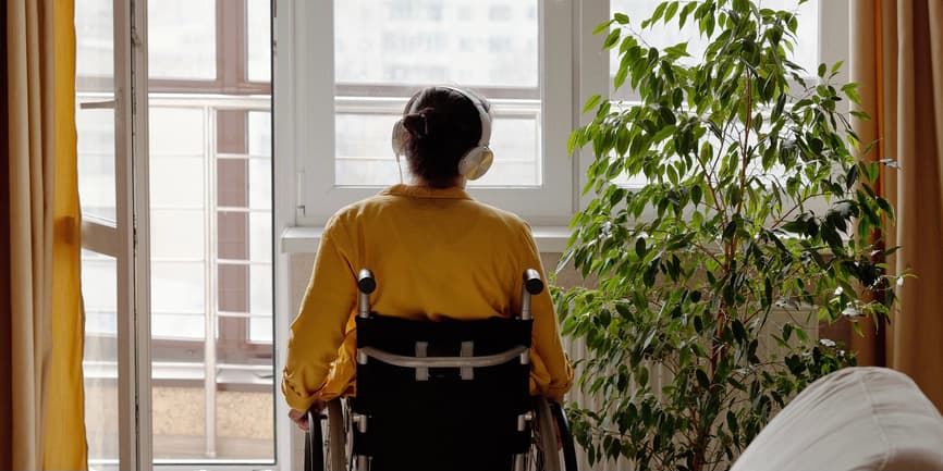 Back view backlit shot of unrecognizable woman in a wheelchair listening to music in her headphones and looking through window thinking of something.