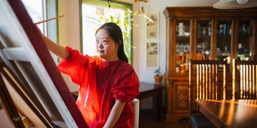 A young Asian woman with Downs syndrome wearing a red dress paints at an easel by windows in a brightly lit, warm dining room.