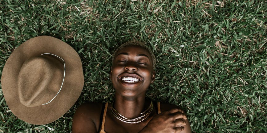 Portrait of a beautiful young Black woman wearing a brown top smiling with her eyes closed as she lays on the grass, with a brown hat next to her.