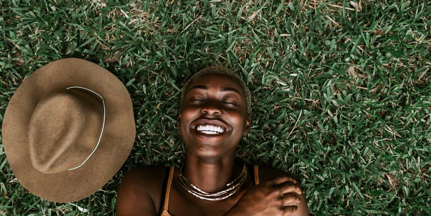 Portrait of a beautiful young Black woman wearing a brown top smiling with her eyes closed as she lays on the grass, with a brown hat next to her.