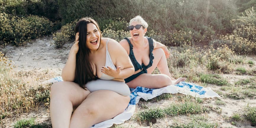 Two white curvy women in bathing suits smile and laugh as they sit on towels on the sandy beach with rays of sunlight coming down through the brush behind them.