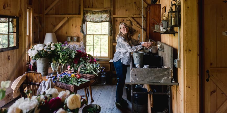 A middle aged white woman with long blond-gray hair arranges flowers to sell in a wooden barn and washes her hands in a sink.