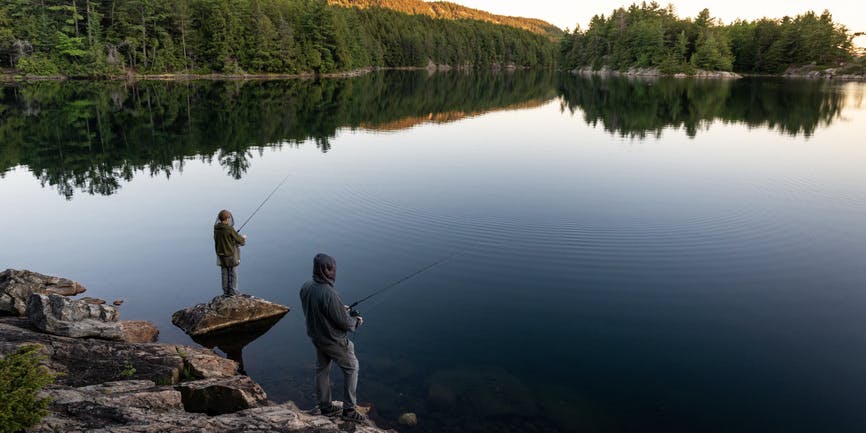 Father and son fishing while on a family backcountry canoe trip near Killarney, Ontario, Canada. Both are wearing mosquito nets over their faces.